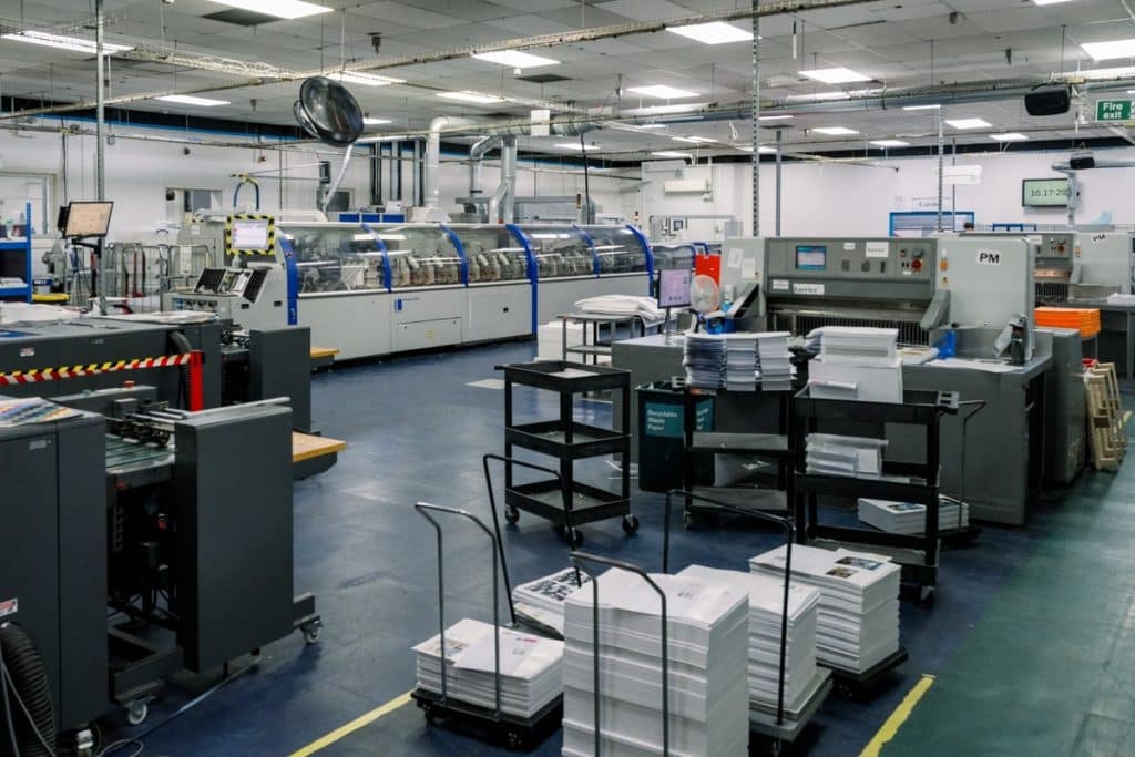 A wide view of Ex Why Zed’s printworks floor with industrial presses and stacks of printed sheets, illustrating litho vs digital printing in action.
