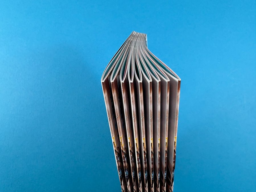 Side view of a stapled saddle-stitched booklet, showing the folded spine and page sections on a blue background.
