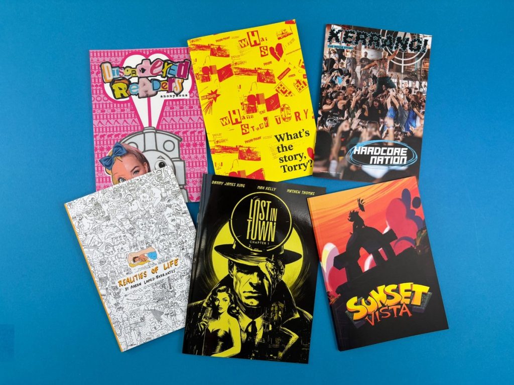 Selection of colourful printed zines and comics laid out on a blue background, showing different formats and cover styles.