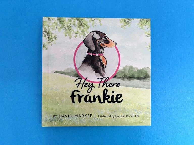 Square children’s hardback cover of a black and tan sausage dog inside a pink ring above a watercolour meadow